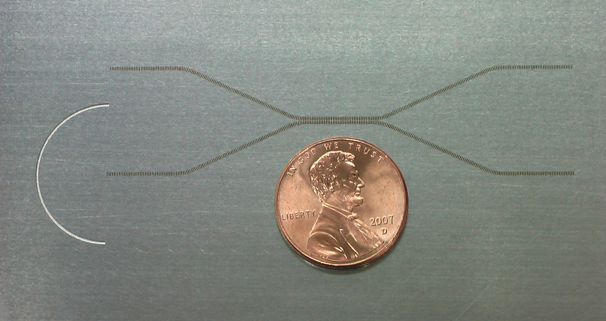 Close-up of a waveguide device that "couples" terahertz radiation, moving it from one wire-like waveguide to another. The device is fabricated on a piece of stainless steel foil. Terahertz radiation is beamed onto the foil within the semicircular etching, which focuses the radiation and sends it down the lower waveguide (the lower part of the "X" shape). Where the two waveguides come near each other (the elongated middle of the "X"), half the radiation jumps from one waveguide to another, so half the radiation comes out of the right-side end of each waveguide. Each waveguide is made of numerous small rectangular punctures in the foil. The penny is for scale.